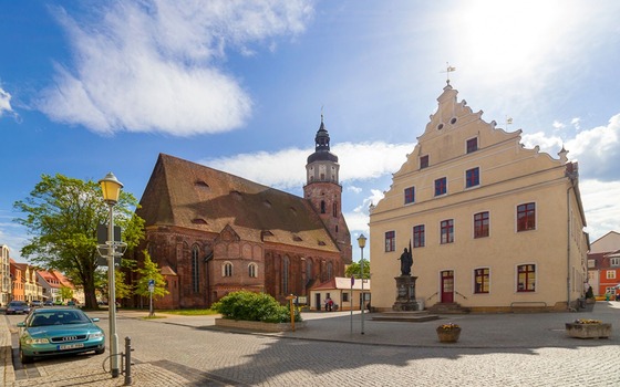 Kopfsteinpflasterplatz mit Backsteinkirche, Kirchturm, hellem Giebelhaus und Statue unter blauem Himmel., Foto: Andreas Franke, Lizenz: Andreas Franke